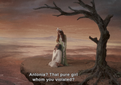 Screenshot from Immortal featuring a woman in a flower-draped dress and crown standing over a bearded man in a white robe kneeling beneath her. They're on an obvious set, dressed as a cliff with a gnarled leafless tree bent over them. She's saying to him "Antonia? That pure girl whom you violated?"