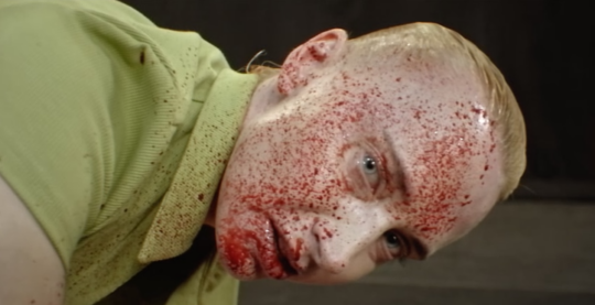 Screenshot from Immortality, where an actor stares into the camera, hair slicked back, face splattered with blood, and a disconcerting look on their face
