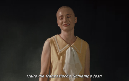 An actor in a large-collared blouse and very close haircut wears a strained smile against a black background. The subtitle is saying something in German I think