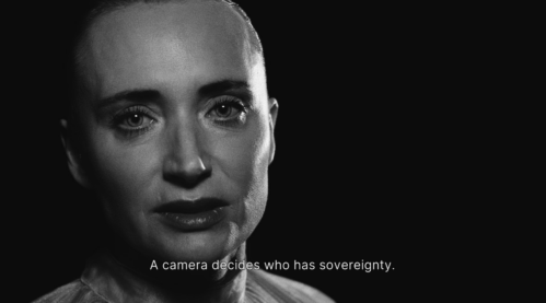 A stark close-up of a short-haired actor, staring into the camera. Subtitle says "A camera decides who has sovereignty."