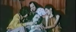 A still from the trailer of Children Shouldn't Play with Dead Things, where the three young adults are dressed in their 70s weirdo finest, in front of some wood paneled walls, caressing a corpse and laughing at their hijinks