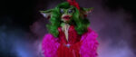 Greta, the female gremlin from Gremlins 2, poses in a sequined red gown and pink feather boa, a large red flower pinned to her shiny green mane of wavy tresses. Her makeup is flawless on her scaly green face.