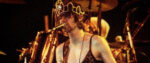 A grainy photograph of Kurt Cobain playing his guitar and singing into a mic while wearing a slip dress and rhinestone tiara.