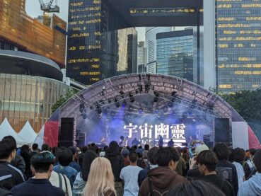 A photo of the band Uchu Yurei playing with some skyscrapers behind them. They're looking down at their instruments as if they're playing some engrossing, challenging music