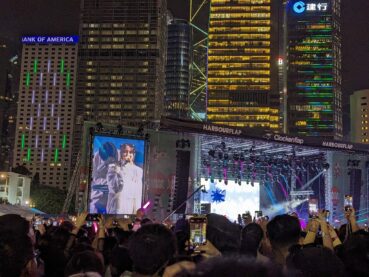 A photo of the band Yoasobi on stage, mostly of the large screen and the towering neon-lit buildings behind the stage. The singer is wearing a stylish outfit and commanding the microphone