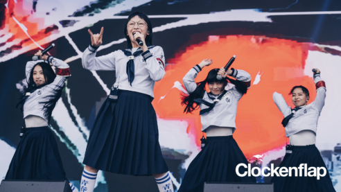 A press photo of the band Atarashii Gakko! performing at Clockenflap in matching student uniforms holding microphones and dancing triumphantly