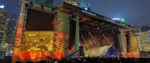 A photo by author Khee Hoon Chan of the main Clockenflap stage, draped on both sides with comic art and behind by the buildings of Hong Kong, and a screen that shows a musician playing hard
