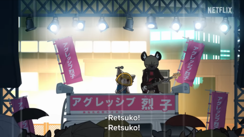 Retsuko and her friend Haida the hyena stand on a dais in front of a crowd with banners in Japanese around them while Haida gets ready to shred a bass and Retsuko holds a microphone triumphantly above her head