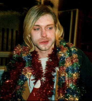 Kurt, adorned in festive multicolored garland, looks just off camera, skeptical and party-worn.