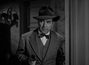 A screenshot from one of the Whistler movies, with a man in a fedora and a bowtie holding a handgun and looking through a room
