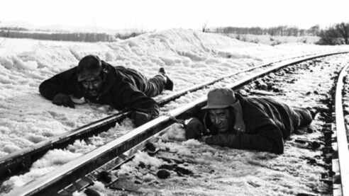 A scene from Abarshiri Prison in black and white, with two prisoners laying prone, one on train tracks and the other in the snow nearby, with handcuffs linking them and the chain on the track