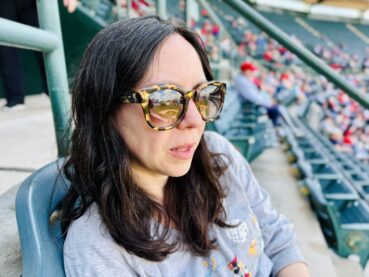 A long-haired brunette woman in chic tortoiseshell sunglasses sits in the plastic molded seats at a baseball stadium, gazing down at the field.