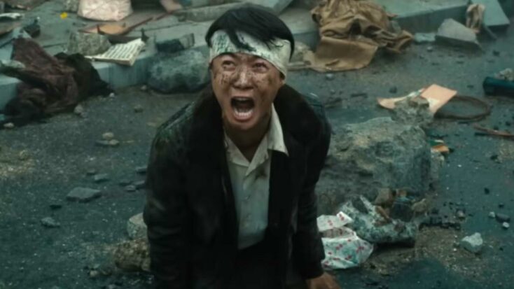 A man in a tattered suit and head bandage kneels wailing on the ground in a still from Godzilla Minus One.