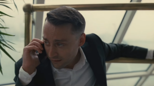 A screenshot from Succession with a young man in suit and white shirt on a boat holding a phone and looking very distressed