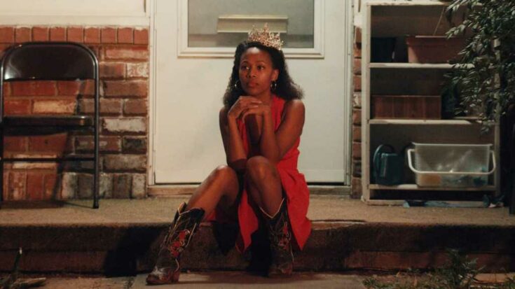 Nicole Behaire sits on her front step dressed in a sleeveless pink dress, cowboy boots, and a tiara in a still from Miss Juneteenth.