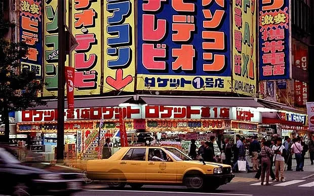 A photograph of Electric City, Akihabara, Tokyo in the the 1970s. Colorful signs reach upwards while a boxy yellow cab sits center of frame.