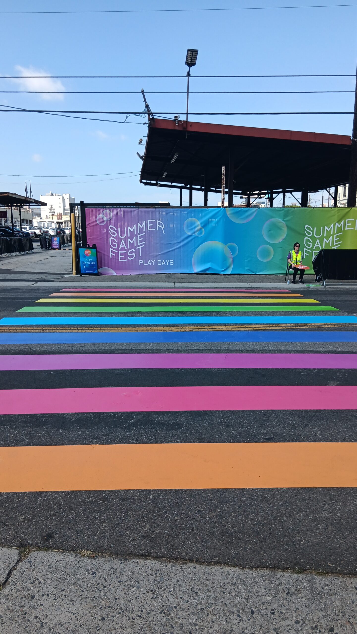 A rainbow crosswalk leads the way to a temporary wall displaying a banner advertising Summer Game Fest's Play Days. A crossing guard in a fluorescent yellow vest sits dutifully on the other side of the road.