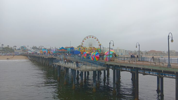 A ferris wheel sits prettily in the distance in a photo of the Santa Monica Pier taken on an overcast day. Rainbow umbrellas lined up along the pier brighten the image considerably.