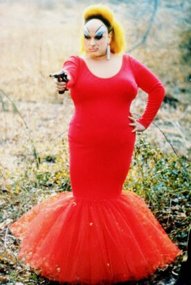 Perhaps the most famous photograph of Divine: In a still from Pink Flamingoes, she stands holding a pistol in a form-fitting red dress, her left hand defiantly on her hip.