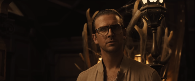 A screenshot from the trailer of Abigail featuring our favorite little guy Dan Stevens in bubbles glasses and a tan bomber jacket and a gold chain looking perplexed but also maybe interested