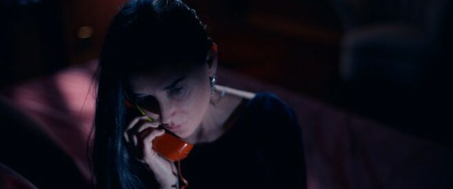 A screenshot from The Substance where Demi Moore sits in a dark room holding a red phone to her face