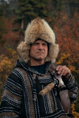 Roland Welker, a contestant on the television show Alone, stands stoically gazing off into the wilderness, his head encased in a massive fur-lined hat.