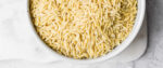 A top-down view of a bowl of uncooked orzo pasta resting on a marble counter.
