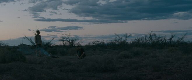 A still from The Rover where two people are searching through a scrubby desert at dusk
