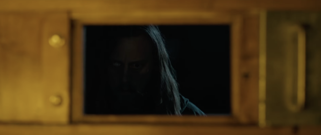 A screenshot from Oddity with an iron door and a small door opened in it to reveal a character with heterochromia mostly hidden in shadow looking at the viewer