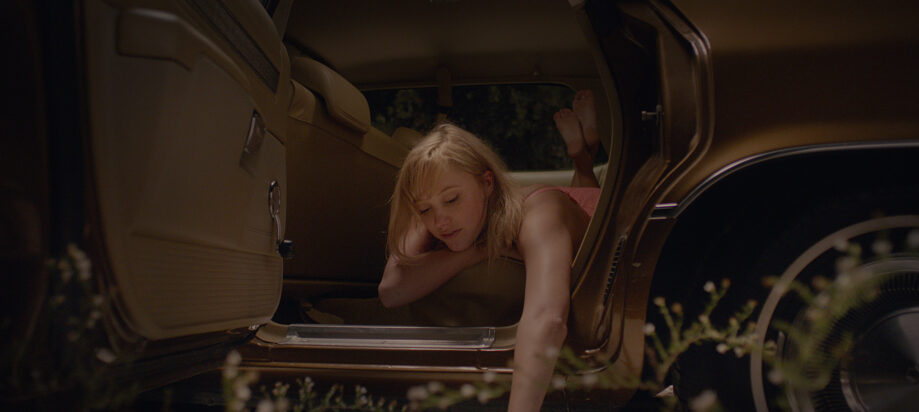 A scene from It Follows shows a young woman lying on her belly in the backseat of a car, trailing her fingers along the ground through the open door.