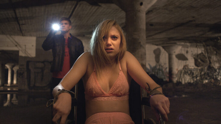 A still from It Follows shows a woman in just her bra tied to a chair while looking worriedly at something offscreen. A man shines a flashlight behind here in the same direction she's looking.