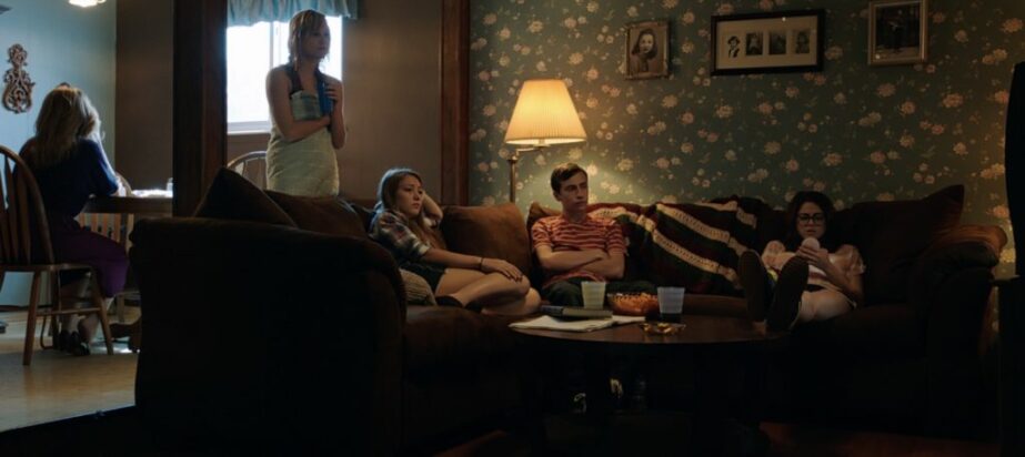 A group of people are gathered in a dimly lit suburban living room, seemingly all focused on a television.