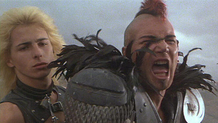 Mohawked Wez and his blonde boy toy sit astride their motorcycle in a still from The Road Warrior.