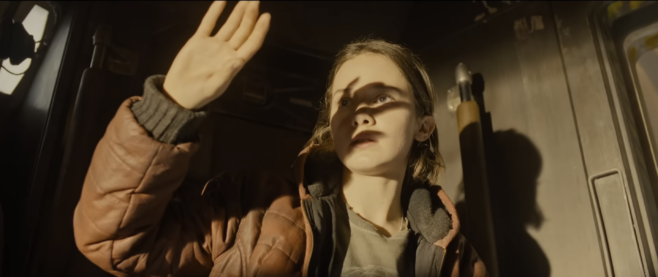 A screenshot from the trailer for Alien Romulus where a worker in a threadbare shirt and puffy jacket holds their hand against a light that is shining over their eyes