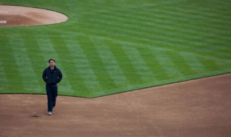 A screenshot from Moneyball where Brad Prit is wearing a track suit and walking across a baseball field with his hands in his pockets and a very serious demeanour