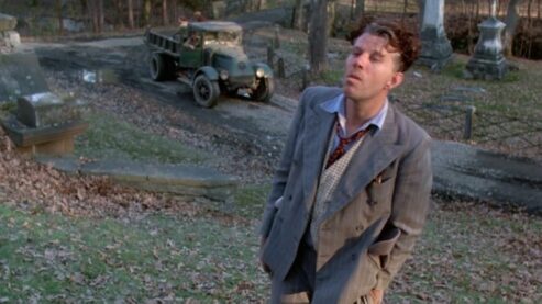 A still from the film Ironweed where Tom Waits is wearing a rumpled suit and standing in a leaf-strewn cemetery as a truck full of dudes unloads in the back