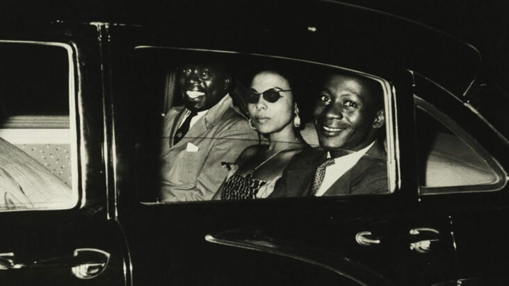 A still from Johan Grimonprez's 'Soundtrack for a Coup d'Etat' shows two men and woman in the backseat of a car, the men smiling at the camera and the woman aloof in dark sunglasses.