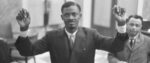 A black-and-white photo of Congolese Prime Minister Patrice Lumumba wearing a suit in what appears to be a government building, arms raised as if in surrender.