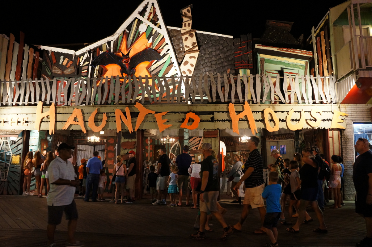 A large crowd gathers around the entrance to a wooden structure at a fair or amusement park, "Haunted House" spelled out in spooky font across the front.