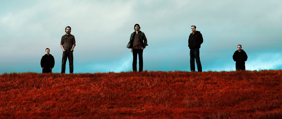 The band Grails stands on the crest of a hill covered in reddish grasses and shrubs, cloudy blue sky behind them.