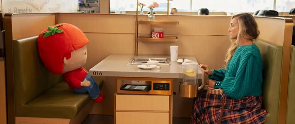 Carrie sits across from a giant stuffed toy with a huge tomato for a head at a Japanese restaurant in a scene from And Just Like That.