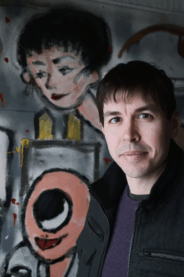 A photograph of Matt Alt with short dark hair and a friendly gaze at the camera with anime inspired graffiti on the wall behind