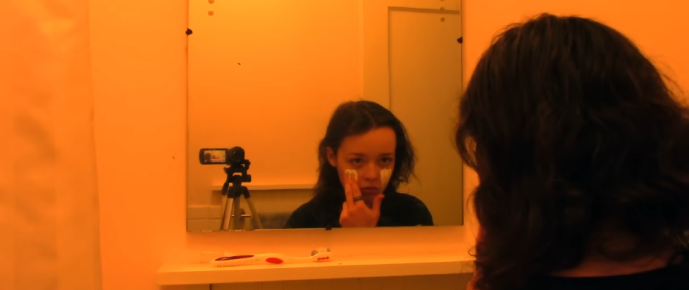 A screenshot from the trailer for We're All Going to the World's Fair where a young person with dark hair in a yellow-lit bathroom is filming themselves putting white liquid on their face