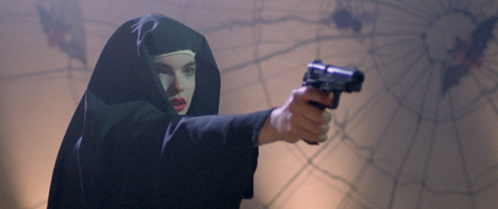 A screenshot from Ms. 45 where a young woman is dressed as a nun and holding a 45 caliber hand gun while standing in front of a spooky spiderweb wall decoration