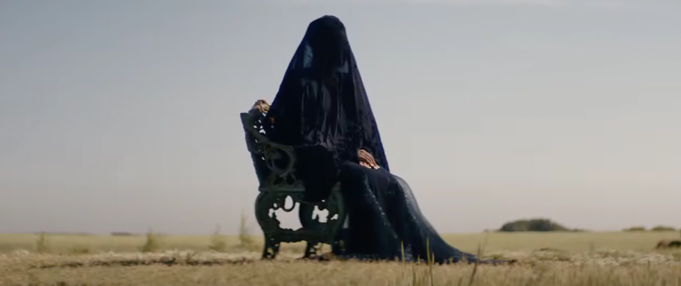 A still from The Woman in the Yard shows a woman draped in black mourning clothes sitting on a chair in a barren yard.