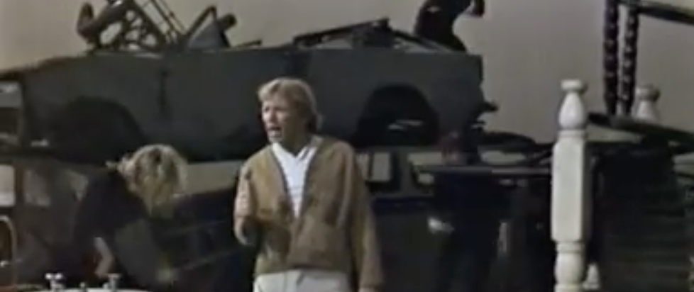 A screenshot of Barry McGuire performing Eve of Destruction on a set decorated with burned out cars and grey wreckage and interpretaive dancers