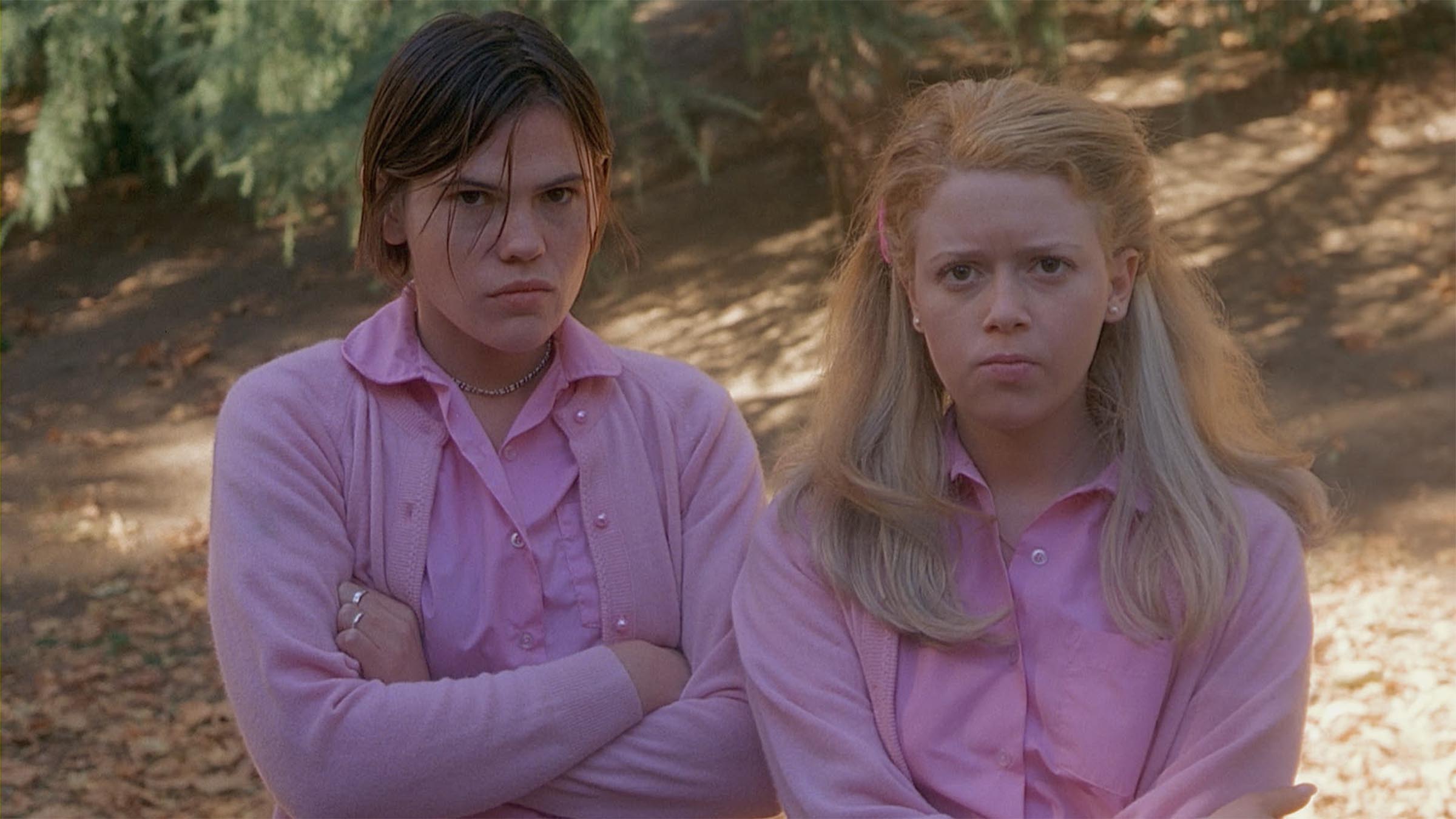 A scene from But I'm a Cheerleader! shows Clea Duvall and Natasha Lyonne's characters standing in the woods looking pissed, awkwardly dressed all in pink.