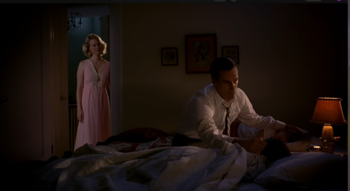 A screencap from Mad Men where Don is sitting next to the bed comforting his son while Betty looks on perhaps misunderstanding Don's feelings at this moment