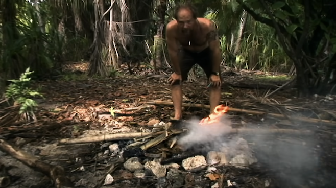 A screenshot from Survivorman where Les is standing in a wooded area over a fire that he just started 