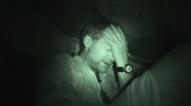 Les Stroud in Survivorman in night vision green holding his head while taking shelter in an abandoned boat for the night